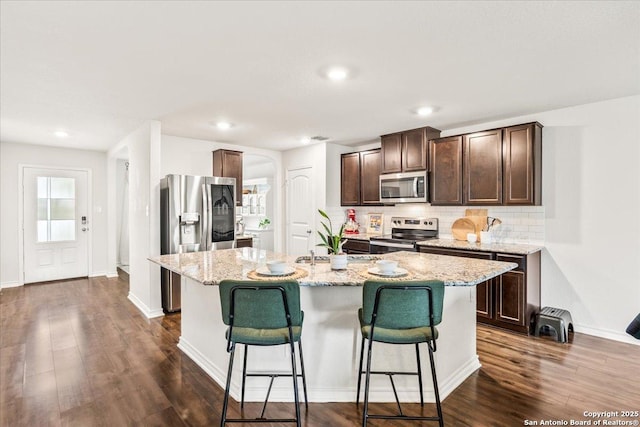 2927 Lindenwood, San Antonio, TX 78245 - kitchen with dark brown cabinetry, backsplash, a center island with sink, stainless steel appliances, and dark wood-type flooring