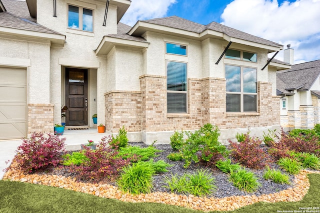 entrance to property featuring brick siding, roof with shingles, and stucco siding