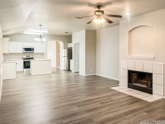 unfurnished living room with light wood finished floors, arched walkways, a tiled fireplace, a ceiling fan, and vaulted ceiling