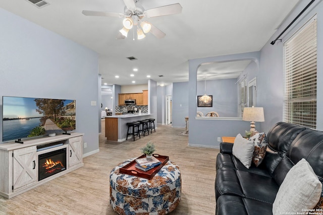 living area featuring a glass covered fireplace, a ceiling fan, baseboards, light wood-type flooring, and visible vents