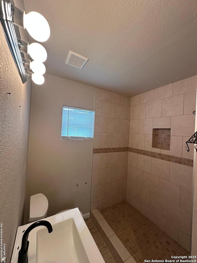 full bath with a sink, toilet, a textured ceiling, visible vents, and tiled shower