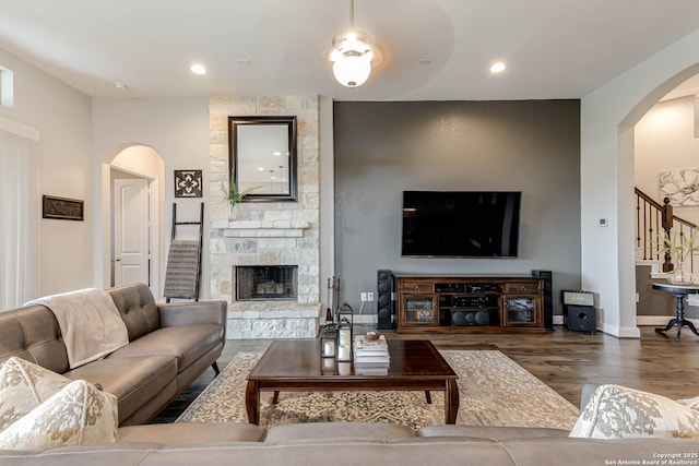 living area featuring arched walkways, a stone fireplace, baseboards, wood finished floors, and stairway