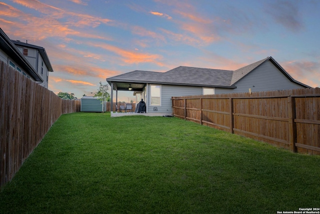 back of property at dusk with a yard, roof with shingles, a fenced backyard, and a patio area