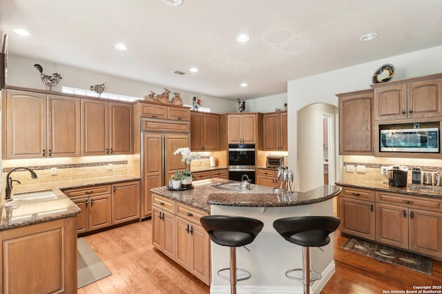 kitchen featuring built in appliances, a kitchen island with sink, tasteful backsplash, a sink, and dark stone countertops