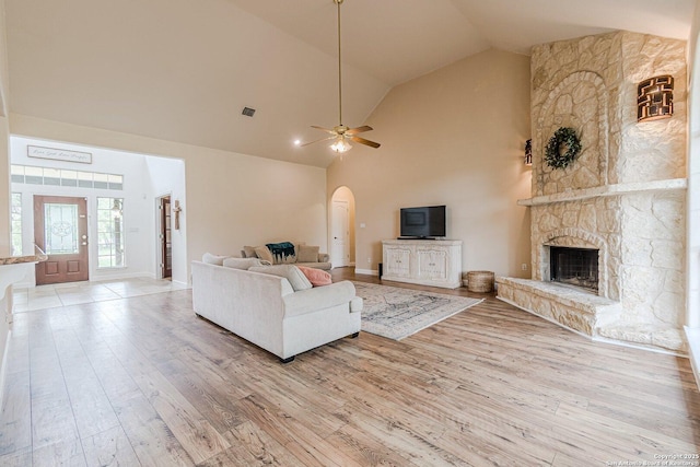 living area featuring a ceiling fan, a stone fireplace, high vaulted ceiling, light wood-type flooring, and arched walkways