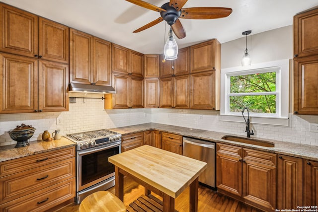 kitchen with a sink, under cabinet range hood, brown cabinets, a ceiling fan, and appliances with stainless steel finishes