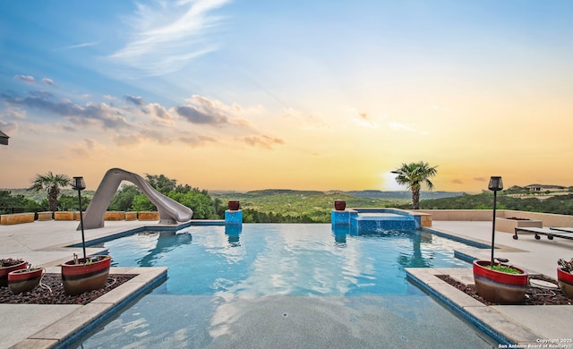 pool at dusk with a patio area, a water slide, and a pool with connected hot tub