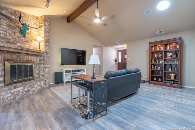 living area with a brick fireplace, a ceiling fan, vaulted ceiling with beams, wood finished floors, and visible vents