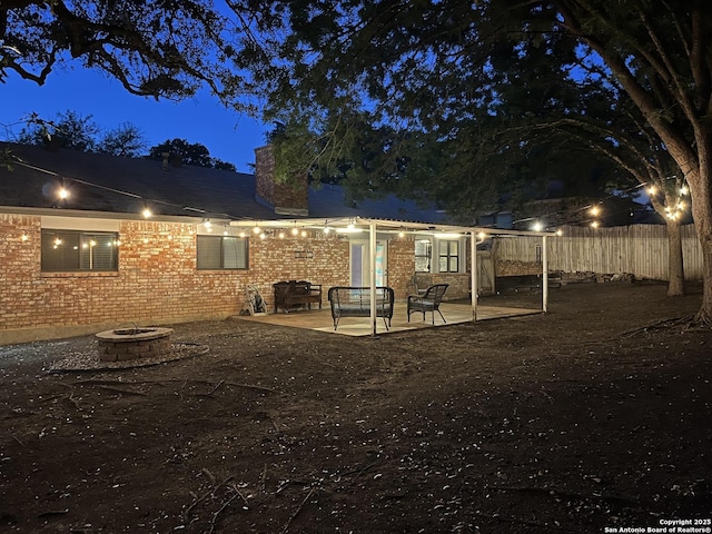back of house at twilight with fence, a chimney, brick siding, a patio area, and an outdoor fire pit