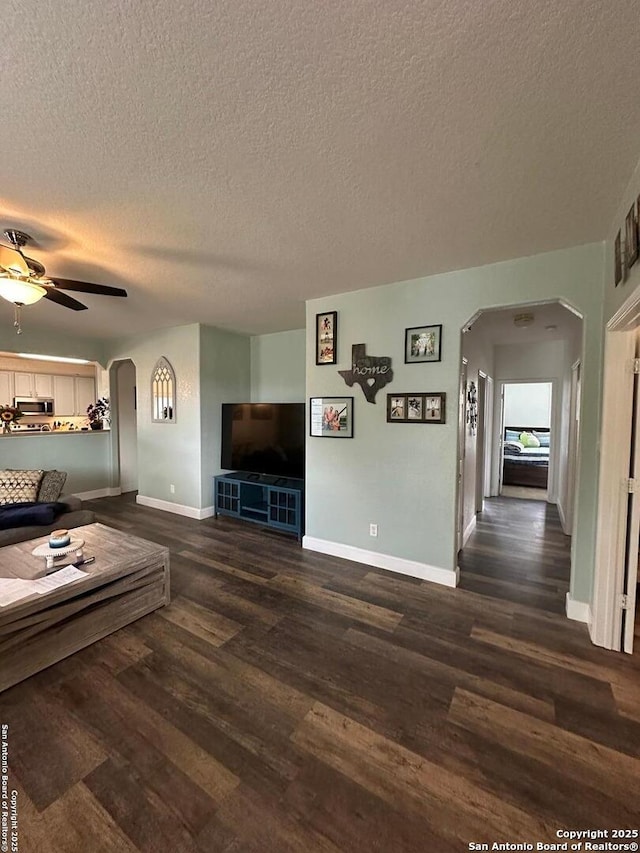 living room featuring ceiling fan, a textured ceiling, arched walkways, baseboards, and dark wood-type flooring