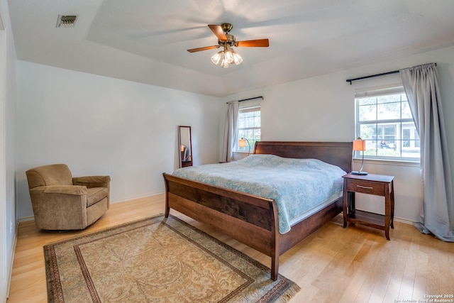 bedroom featuring visible vents, baseboards, ceiling fan, a tray ceiling, and light wood-style flooring