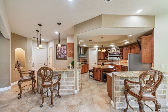 kitchen with wall chimney range hood, a kitchen breakfast bar, brown cabinetry, freestanding refrigerator, and a kitchen island