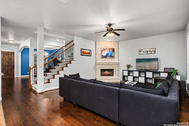 living area featuring a stone fireplace, a ceiling fan, baseboards, dark wood-style floors, and stairway