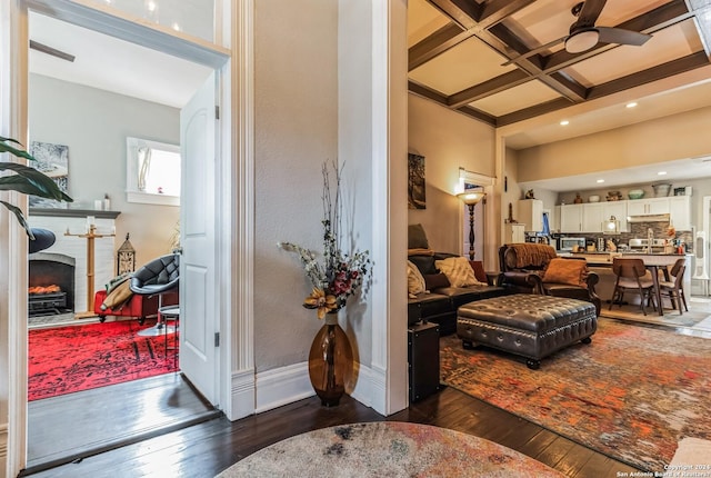 living area with coffered ceiling, beam ceiling, a brick fireplace, hardwood / wood-style floors, and ceiling fan