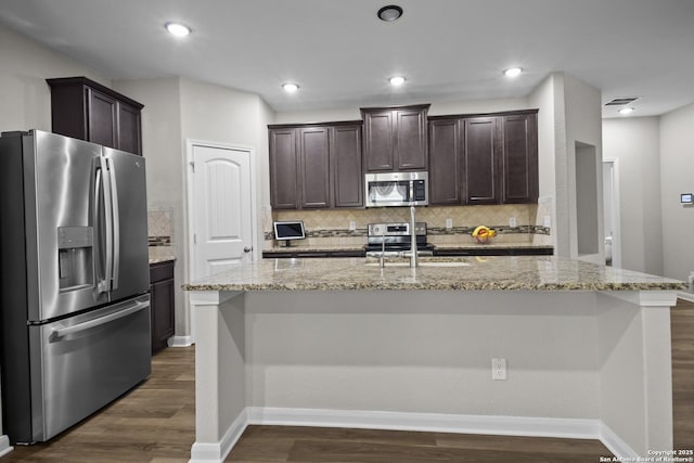kitchen with stainless steel appliances, tasteful backsplash, a kitchen island with sink, dark wood-style floors, and dark brown cabinets