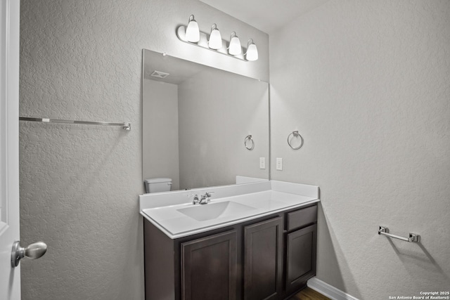bathroom with toilet, visible vents, vanity, and a textured wall