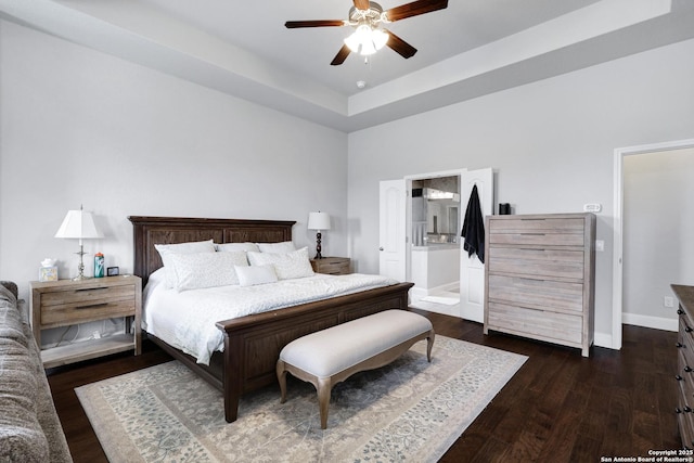 bedroom featuring wood finished floors, a ceiling fan, a tray ceiling, baseboards, and ensuite bath