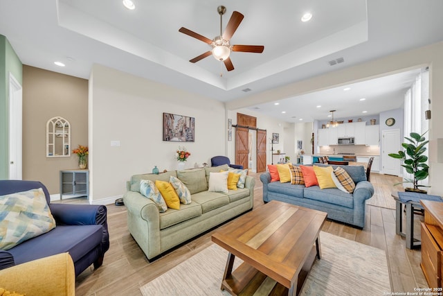 living room with a tray ceiling, light wood-type flooring, a barn door, and a ceiling fan
