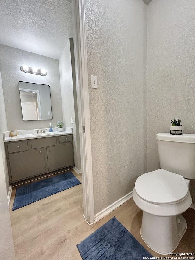 half bath featuring toilet, wood finished floors, vanity, a textured wall, and baseboards