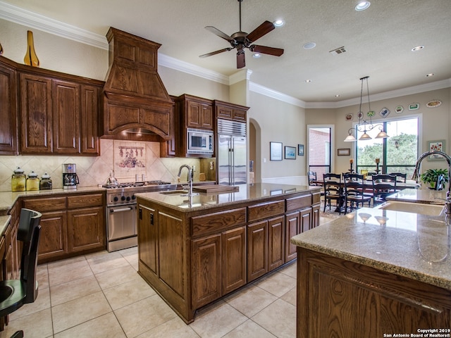 kitchen with built in appliances, a ceiling fan, custom exhaust hood, a sink, and a center island with sink