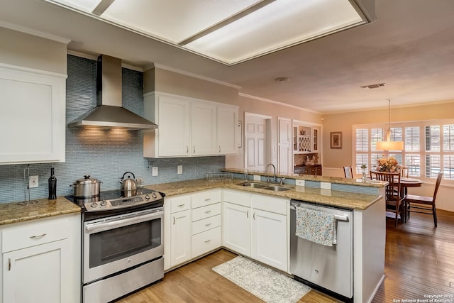 kitchen featuring stainless steel appliances, wall chimney range hood, a sink, a peninsula, and light wood-style floors