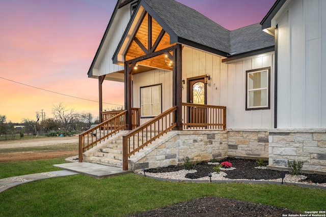 exterior entry at dusk featuring a yard, a shingled roof, stone siding, and board and batten siding
