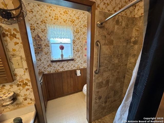 full bathroom with a tile shower, toilet, wallpapered walls, and a wainscoted wall