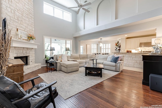 living room featuring ceiling fan, a high ceiling, a stone fireplace, and dark wood-style flooring