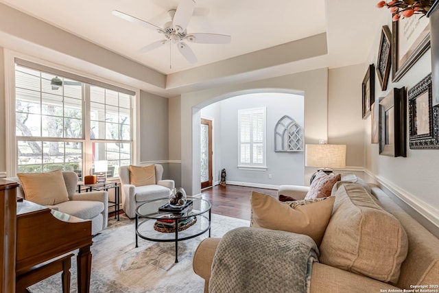 living area featuring wood finished floors, a ceiling fan, a tray ceiling, baseboards, and arched walkways
