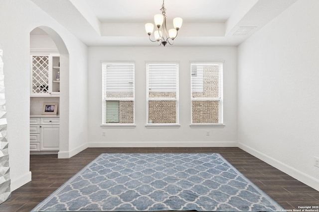 unfurnished dining area featuring a tray ceiling, dark wood-type flooring, a chandelier, and baseboards