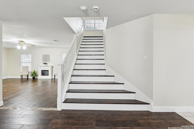staircase featuring ceiling fan, a glass covered fireplace, wood finished floors, and baseboards
