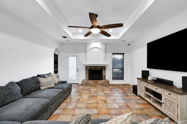 living room featuring a tray ceiling, a tile fireplace, a ceiling fan, plenty of natural light, and crown molding