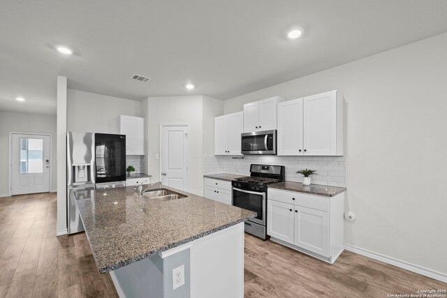 kitchen with appliances with stainless steel finishes, a sink, an island with sink, light wood-type flooring, and dark stone counters