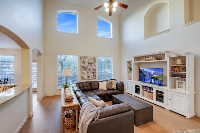 living room featuring arched walkways, ceiling fan, light wood-style flooring, a towering ceiling, and baseboards