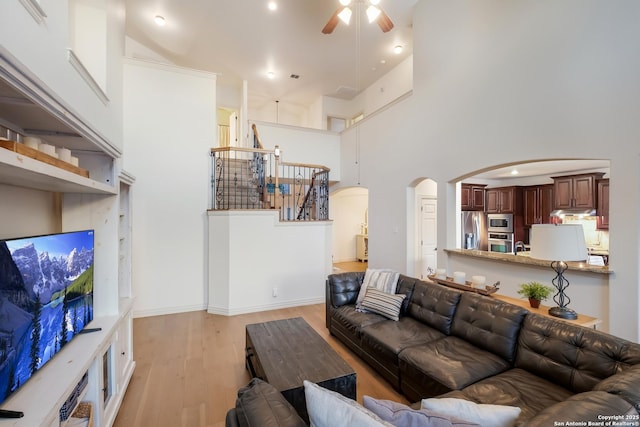 living area featuring arched walkways, a ceiling fan, light wood-style floors, a high ceiling, and stairway