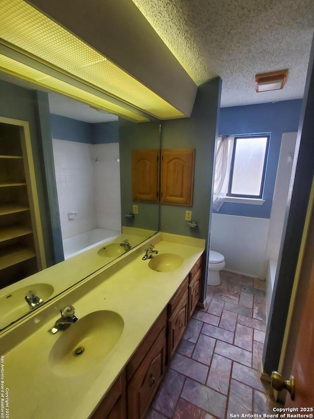 bathroom with stone tile flooring, a bath, toilet, double vanity, and a textured ceiling