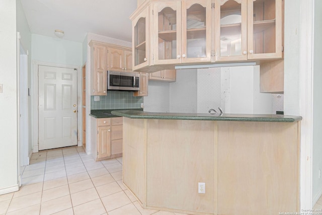 kitchen with stainless steel microwave, light tile patterned floors, light brown cabinetry, backsplash, and glass insert cabinets