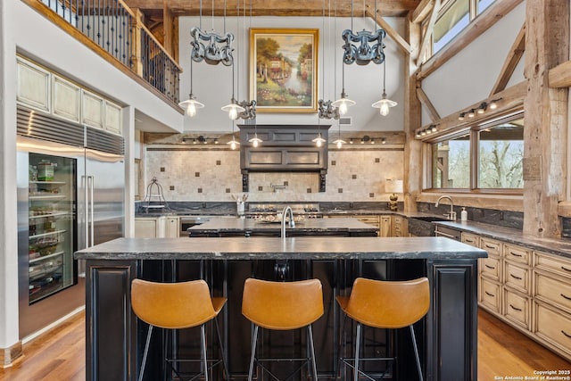 kitchen with dark countertops, a sink, a kitchen island with sink, cream cabinetry, and tasteful backsplash