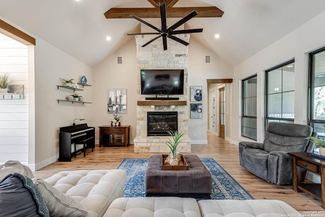 living room with ceiling fan, beam ceiling, a stone fireplace, high vaulted ceiling, and light wood-style flooring