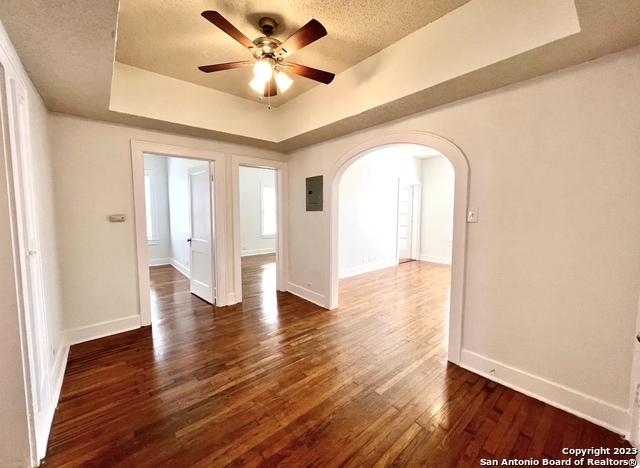 unfurnished room with arched walkways, ceiling fan, dark wood-style floors, a tray ceiling, and baseboards