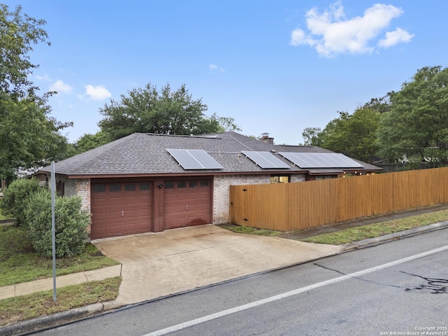 view of front facade with a shingled roof, brick siding, roof mounted solar panels, a garage, and fence