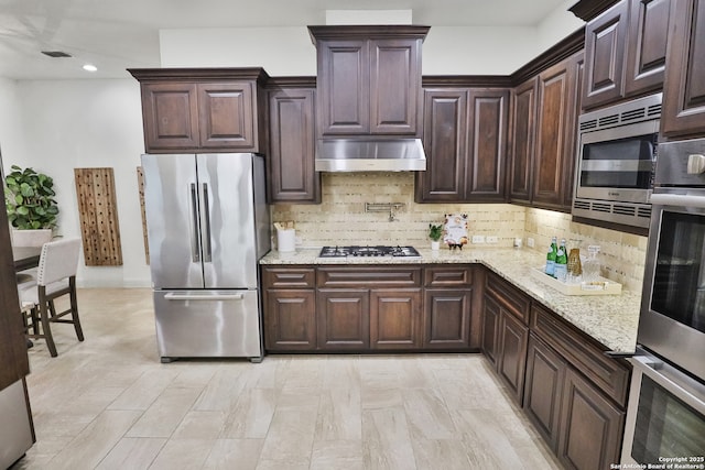 kitchen featuring appliances with stainless steel finishes, under cabinet range hood, decorative backsplash, light stone countertops, and dark brown cabinets