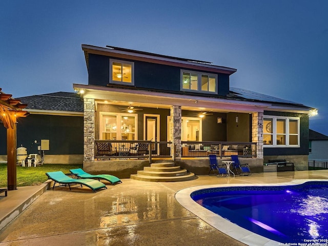back of house at night with an outdoor pool, roof mounted solar panels, a patio, stone siding, and a porch