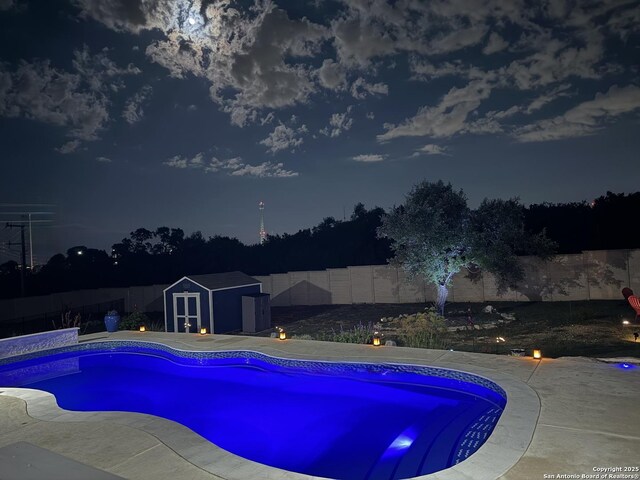 pool at night with a storage shed, a fenced backyard, and an outdoor structure