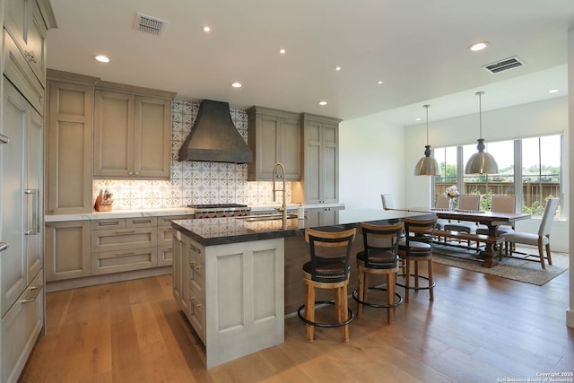 kitchen with custom exhaust hood, a sink, backsplash, light wood-style floors, and an island with sink
