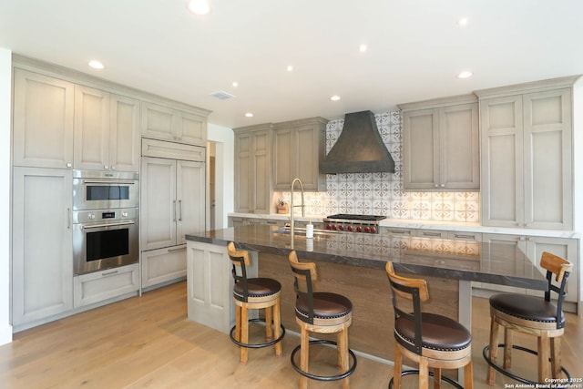 kitchen featuring custom exhaust hood, a sink, stainless steel appliances, backsplash, and light wood-type flooring