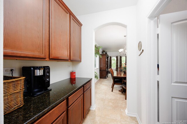 kitchen featuring arched walkways, dark stone counters, light tile patterned flooring, brown cabinetry, and baseboards
