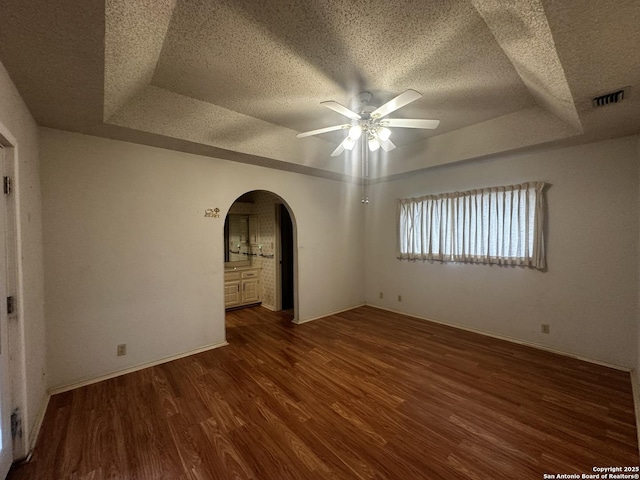 unfurnished room featuring a raised ceiling, a textured ceiling, arched walkways, ceiling fan, and dark wood-style flooring