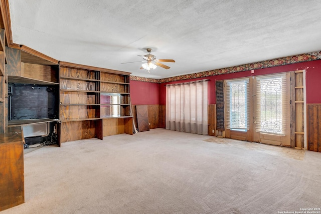 unfurnished living room with a wainscoted wall, carpet, a textured ceiling, a ceiling fan, and wood walls