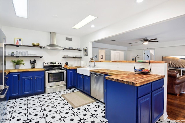 kitchen featuring wood counters, wall chimney range hood, stainless steel appliances, blue cabinetry, and open shelves